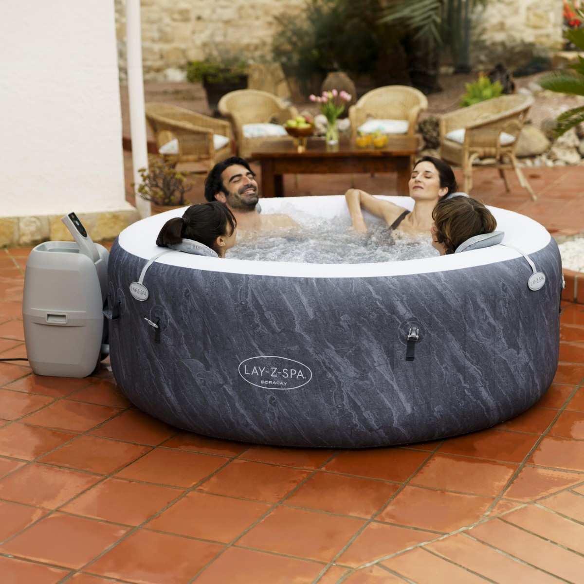 Zestaw jauzzi Lay-Z-Spa Boracay w użyciu: cztery osoby relaksują się w okrągłym dmuchanym jacuzzi ogrodowym z włączonym hydromasażem, obok widoczna pompa filtrująco-grzewcza, jacuzzi ustawione na tarasie w ogrodzie.